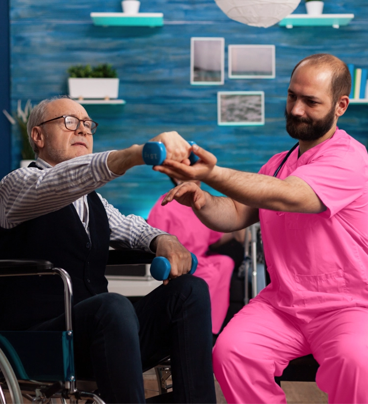 A caregiver in pink scrubs assists a senior man in a wheelchair with physical therapy, helping him lift small blue dumbbells.