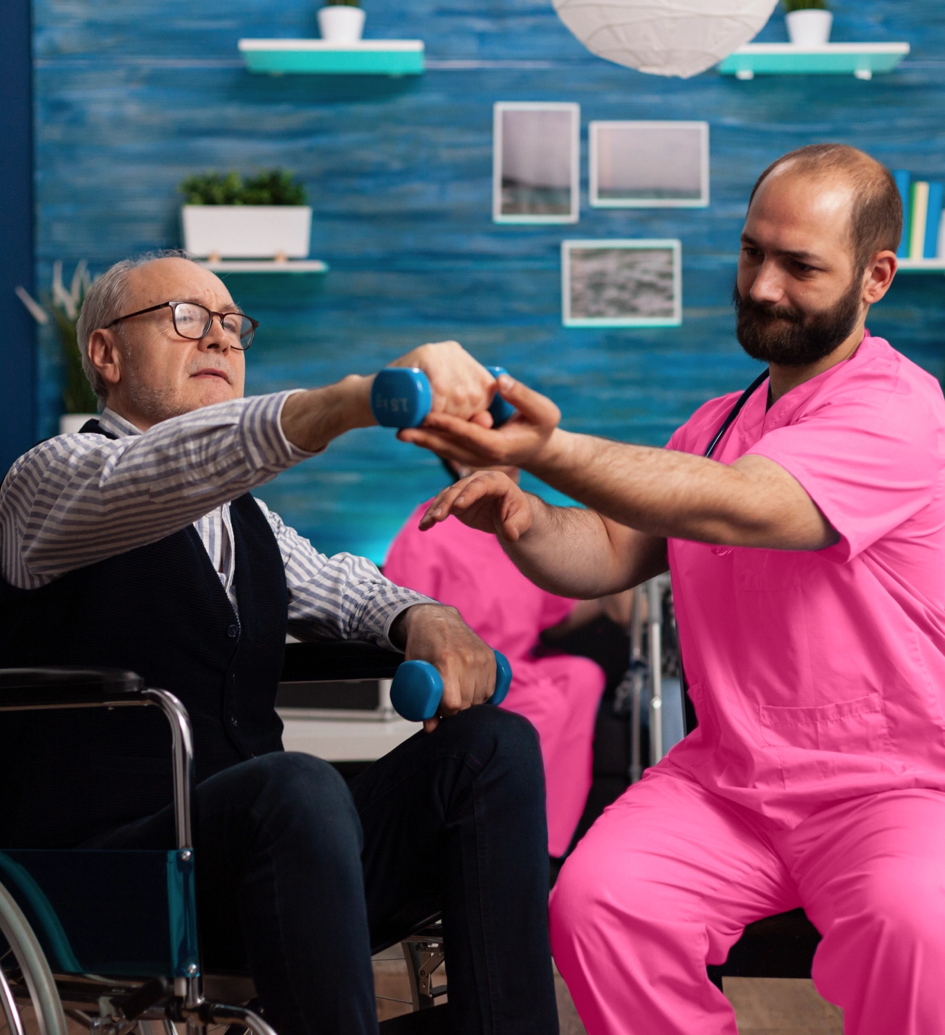 A caregiver in pink scrubs assists a senior man in a wheelchair with physical therapy, helping him lift small blue dumbbells.
