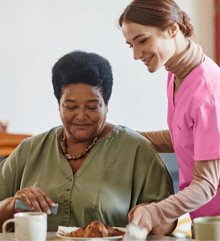 A smiling caregiver in a pink uniform stands behind an older woman who is eating, placing a dish on the table.